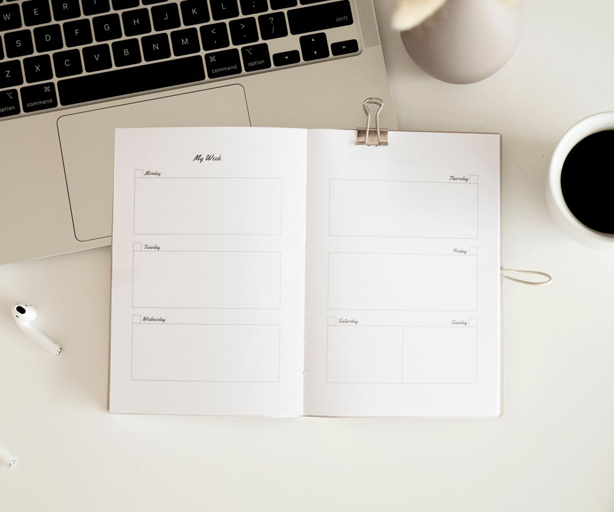 An open weekly planner with a laptop, earbuds and coffee on a light desk.