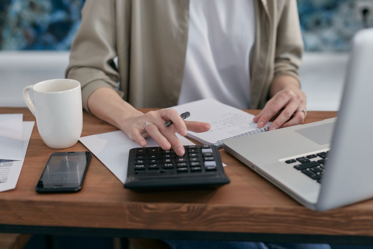 A person checking figures with a calculator and notebook beside a laptop.
