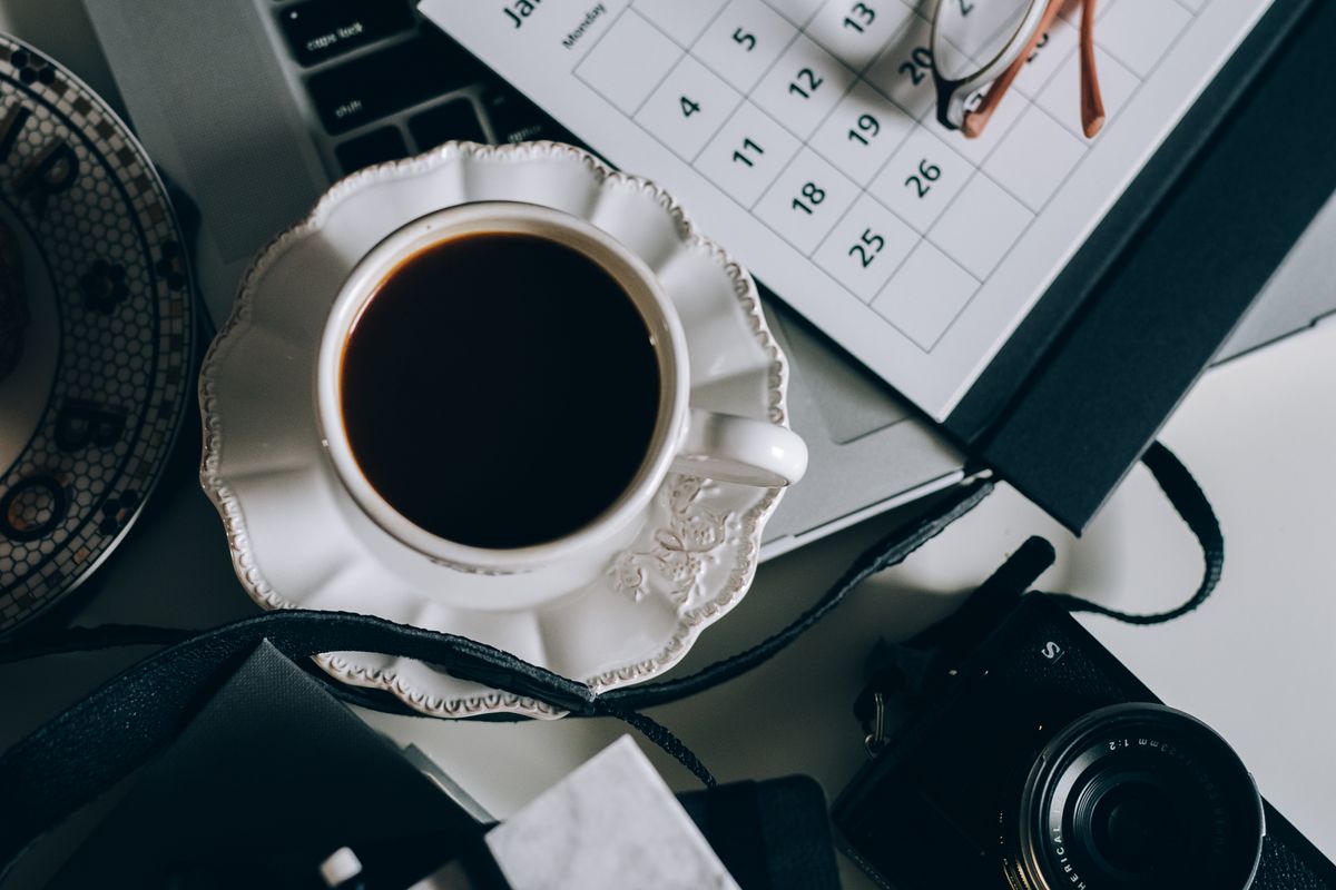 A cup of black coffee next to a calendar, camera and laptop on a desk.